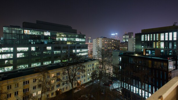 Un paisaje urbano nocturno con edificios modernos, incluyendo un rascacielos con ventanas iluminadas y un edificio más bajo con planta baja visible.