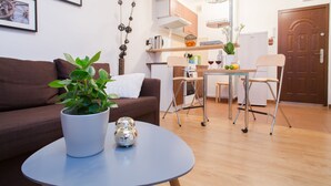 Una sala moderna con un sofá marrón, una mesa de centro redonda con una planta en maceta y esferas decorativas, un comedor con mesa y sillas, y una cocina al fondo.