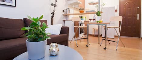 Una sala moderna con un sofá marrón, una mesa de centro redonda con una planta en maceta y esferas decorativas, un comedor con mesa y sillas, y una cocina al fondo.
