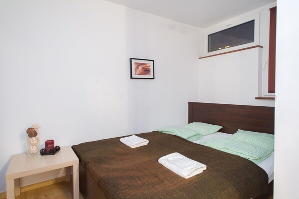 A neatly made bed with a brown bedspread, a green and white patterned blanket, and two folded white towels on a wooden nightstand beside it. - ShortStayPoland Graniczna (Warsaw)