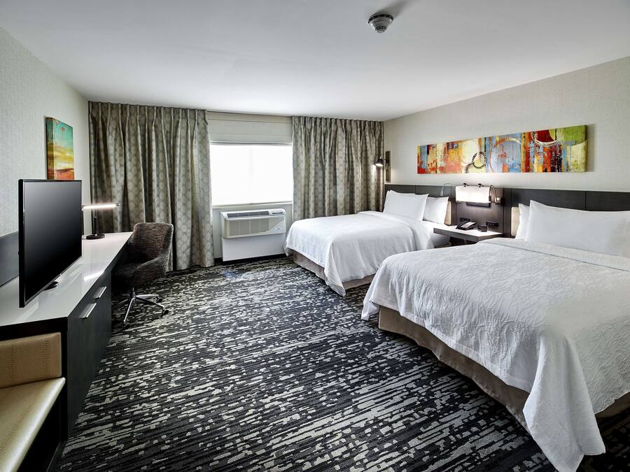 A hotel room with two beds, a flat-screen TV, a desk with a chair, a window with curtains, and a painting on the wall.