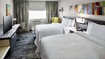 Quarto de hotel com duas camas, uma escrivaninha com computador, uma cadeira e uma janela grande com cortinas.