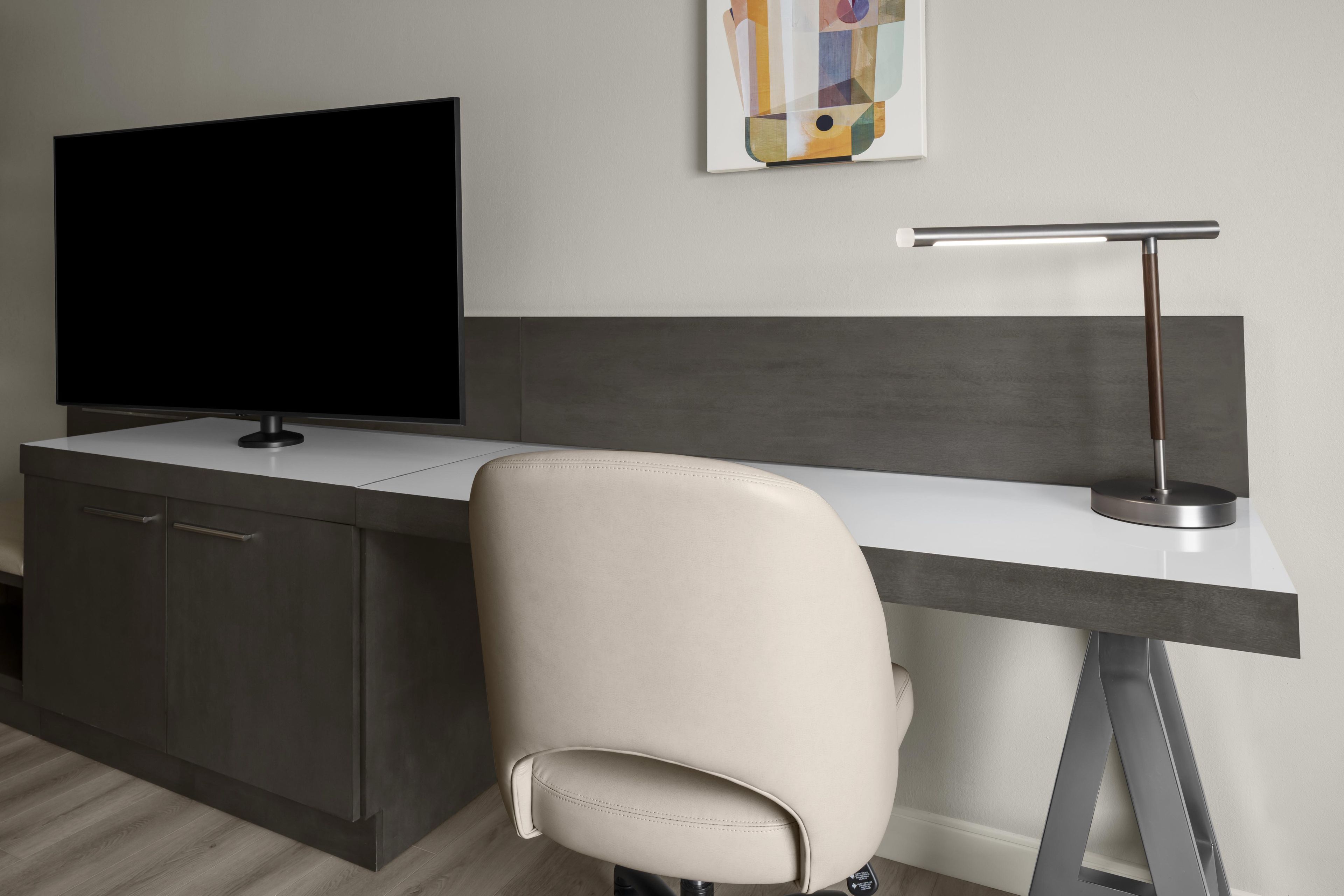 A modern office space with a flat-screen TV, a desk, a chair, and a wall-mounted lamp.