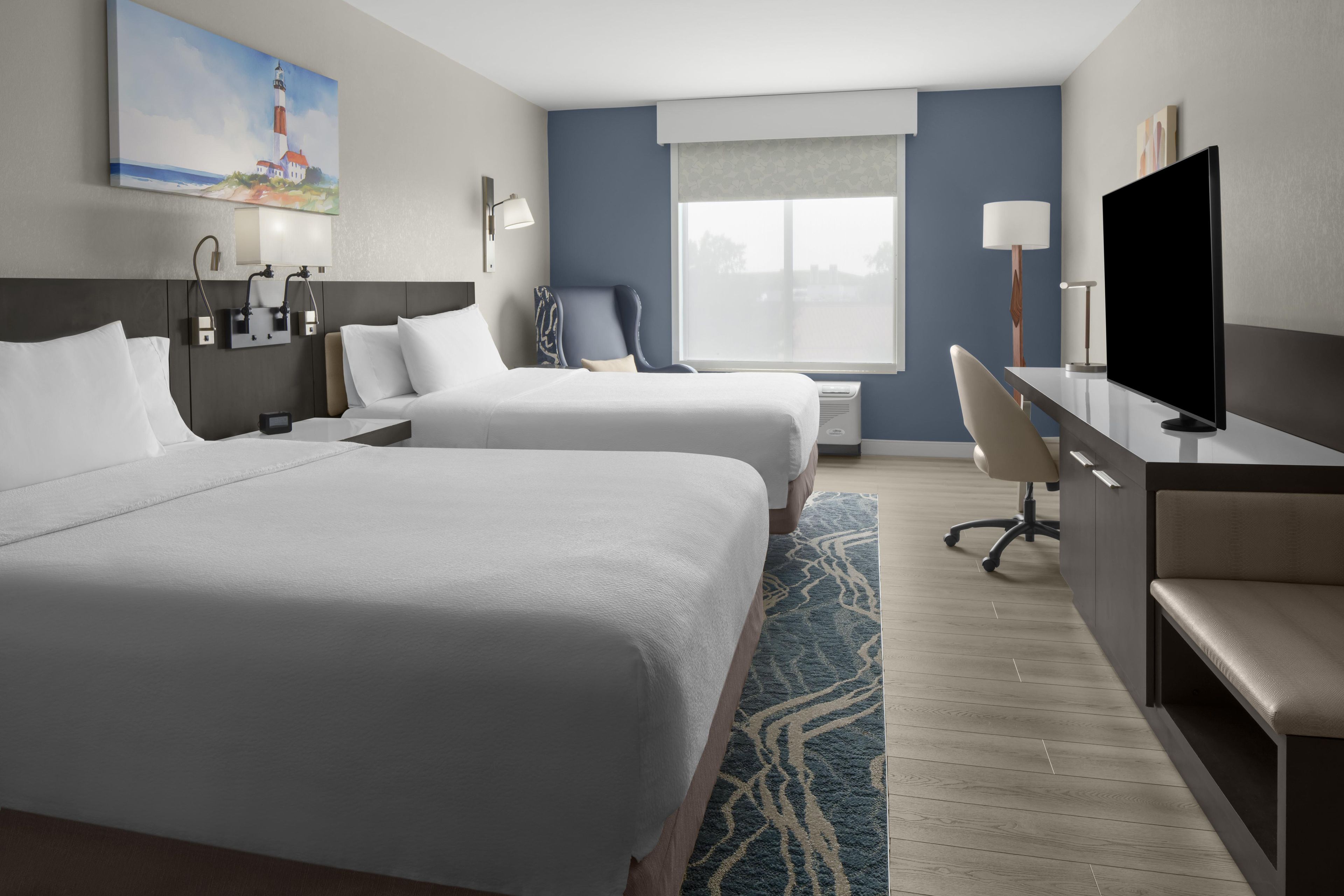 A hotel room with two beds, a desk, a chair, a television, and a painting on the wall.