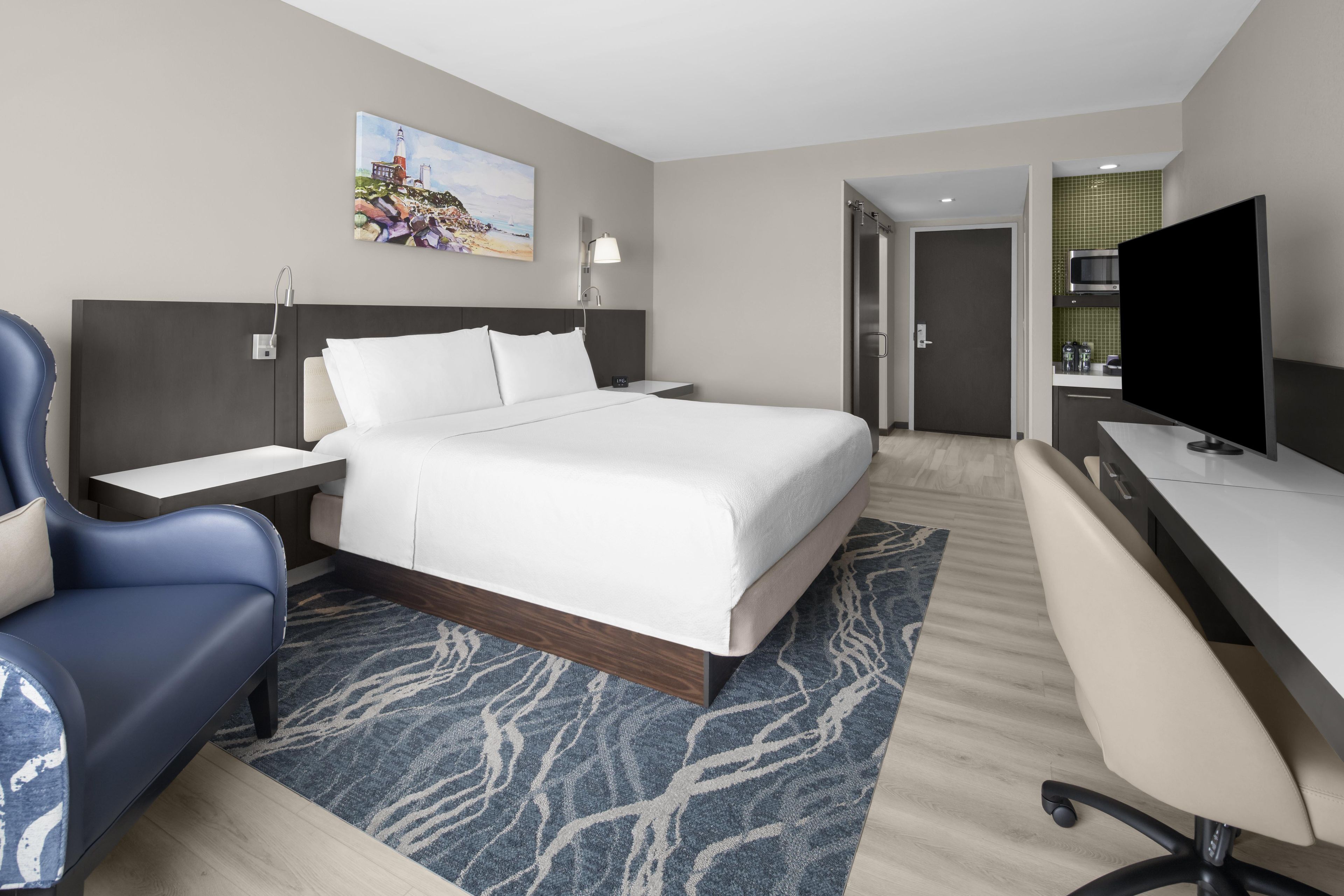 A hotel room with a bed, a desk, a chair, a television, and a painting on the wall.