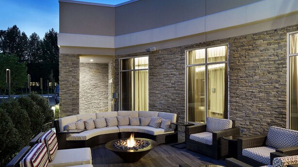 A modern outdoor living area with a curved sofa, wicker furniture, and a central fire pit. - Hilton Garden Inn Roslyn (Port Washington)