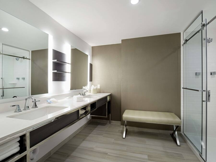 A modern bathroom with a large mirror, two sinks, a bench, and a shower area.