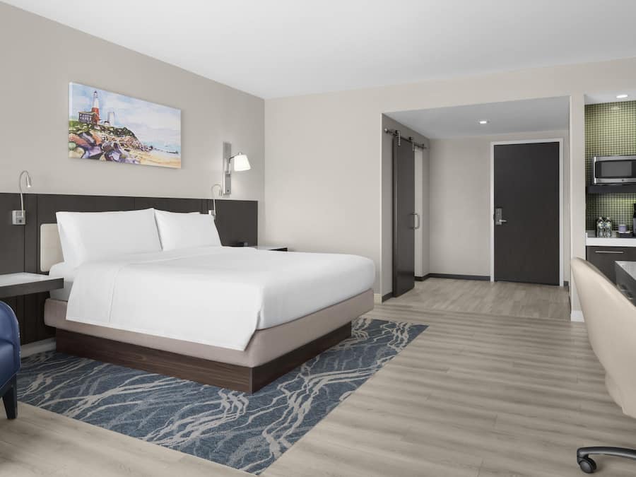 A modern hotel room with a bed, a desk, a chair, a TV, and a painting on the wall.