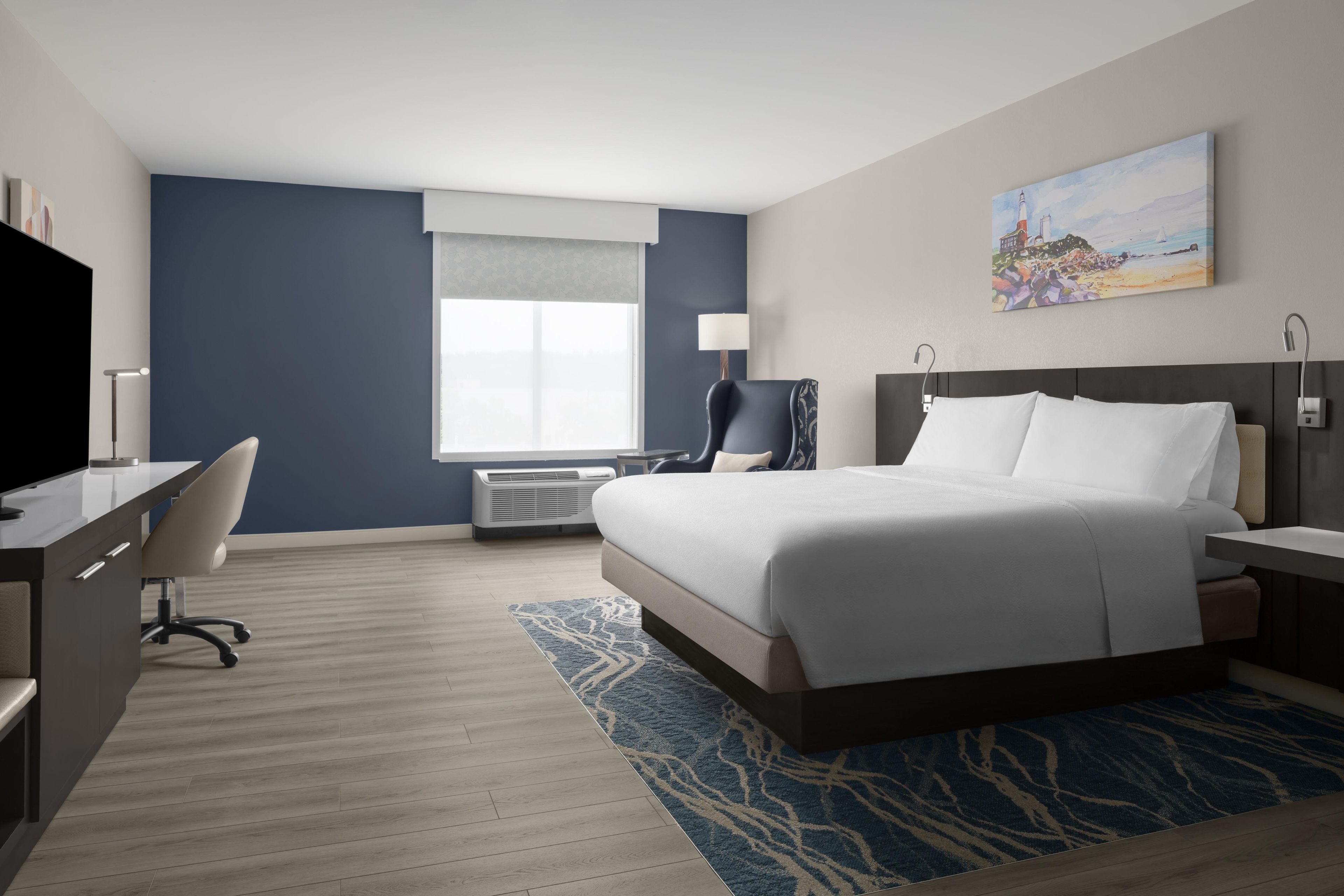A hotel room with a bed, desk, chair, and a painting on the wall.