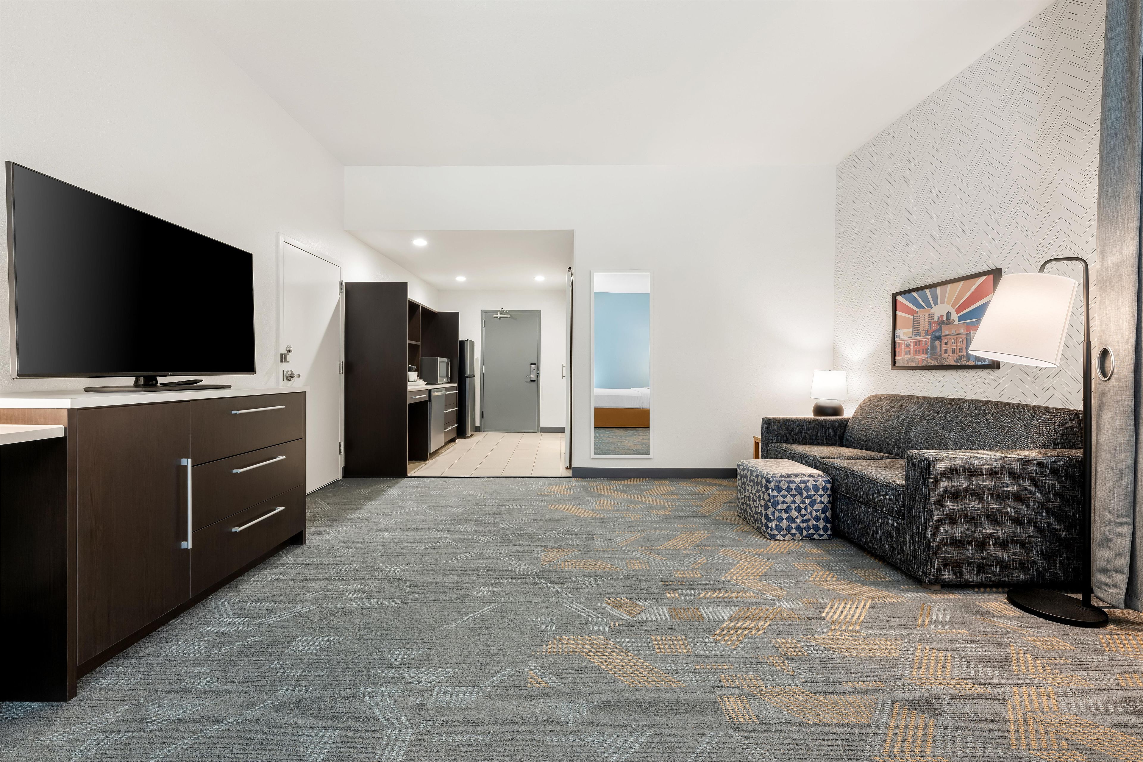 A hotel room with a flat-screen TV, a sofa, a patterned carpet, and a hallway leading to a bathroom.