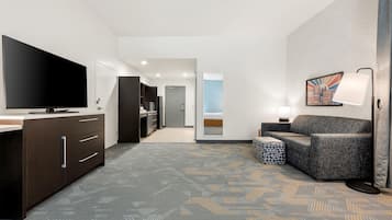 A hotel room with a flat-screen TV, a sofa, a patterned carpet, and a hallway leading to a bathroom.