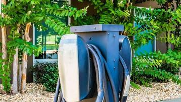 A charging station with multiple cables, set against a backdrop of greenery and a building.