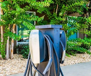 A charging station with multiple cables, set against a backdrop of greenery and a building. - Home2 Suites by Hilton Prattville (Prattville)