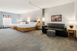 A hotel room with a bed, a sofa, a patterned carpet, and a cityscape painting on the wall. - Home2 Suites by Hilton Prattville (Prattville)