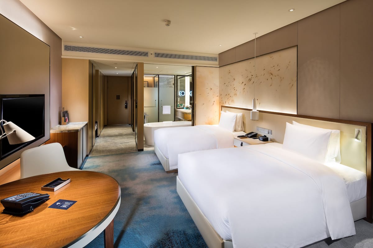 A hotel room with two beds, a desk with a chair, a TV, and a bathroom visible through an open door.
