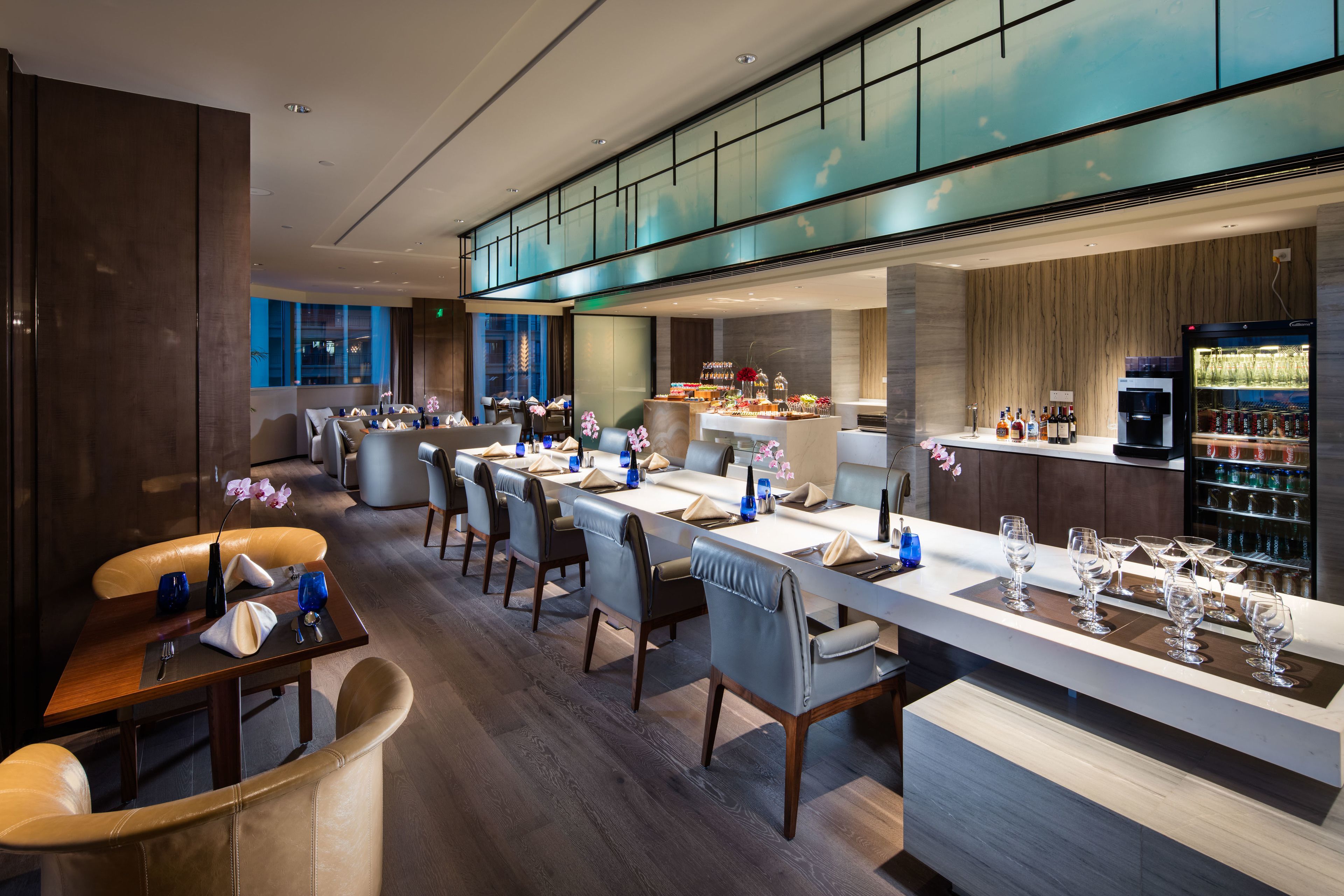 A modern restaurant with a long dining table, multiple chairs, and a bar area with glasses and bottles.
