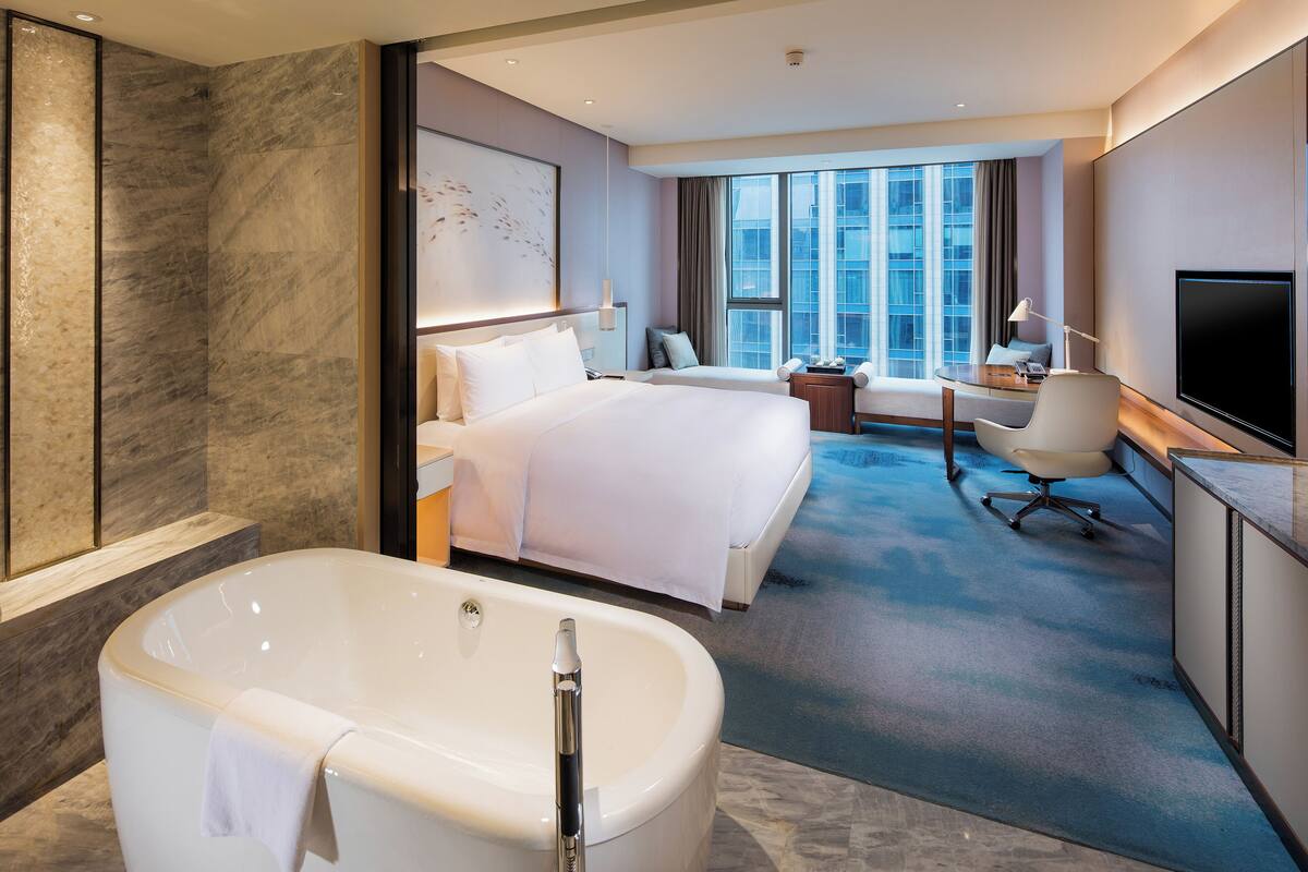 A modern hotel room with a large bed, a separate sitting area, a desk, a flat-screen TV, and a large bathtub.