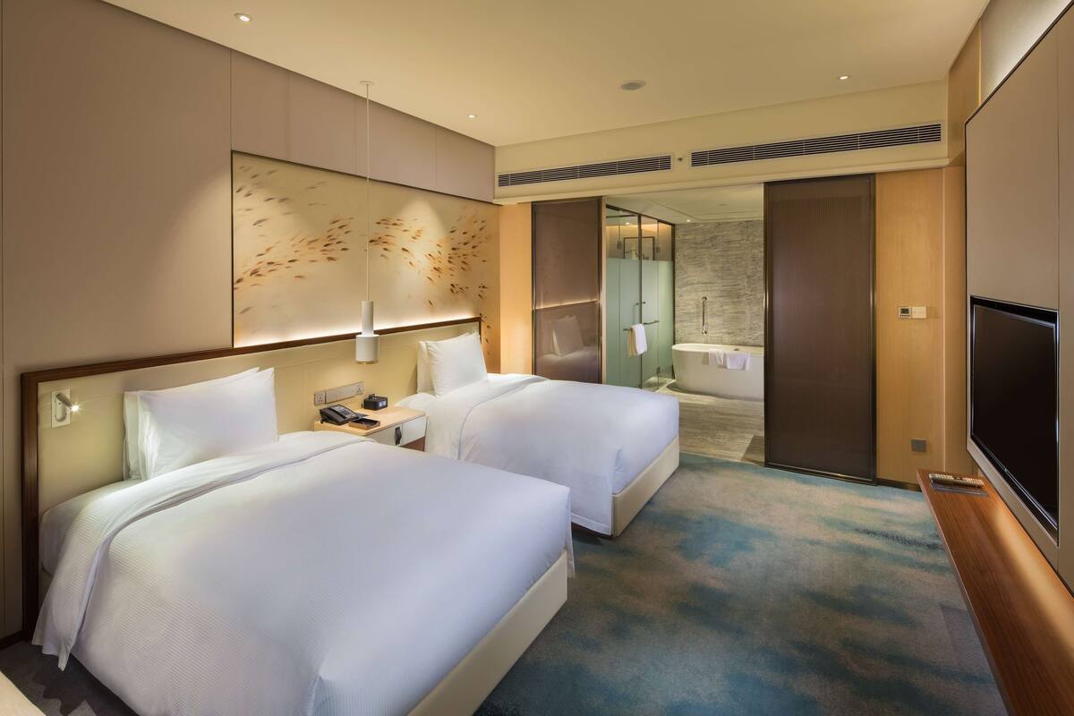 A hotel room with two beds, a TV, a desk, and a bathroom visible through an open door.