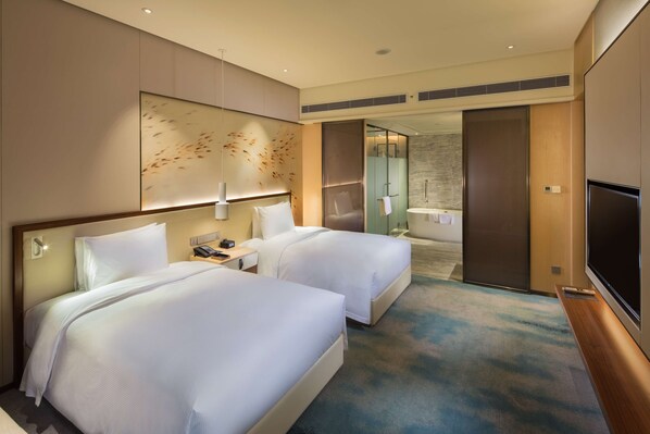 A hotel room with two beds, a TV, a desk, and a bathroom visible through an open door.