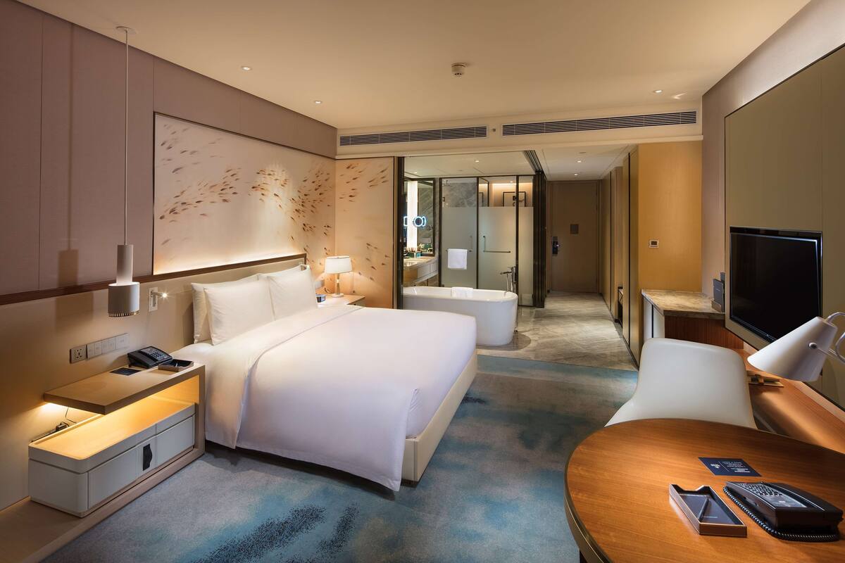 A hotel room with a large bed, bedside tables, a desk, a TV, and a bathroom visible through an open door.
