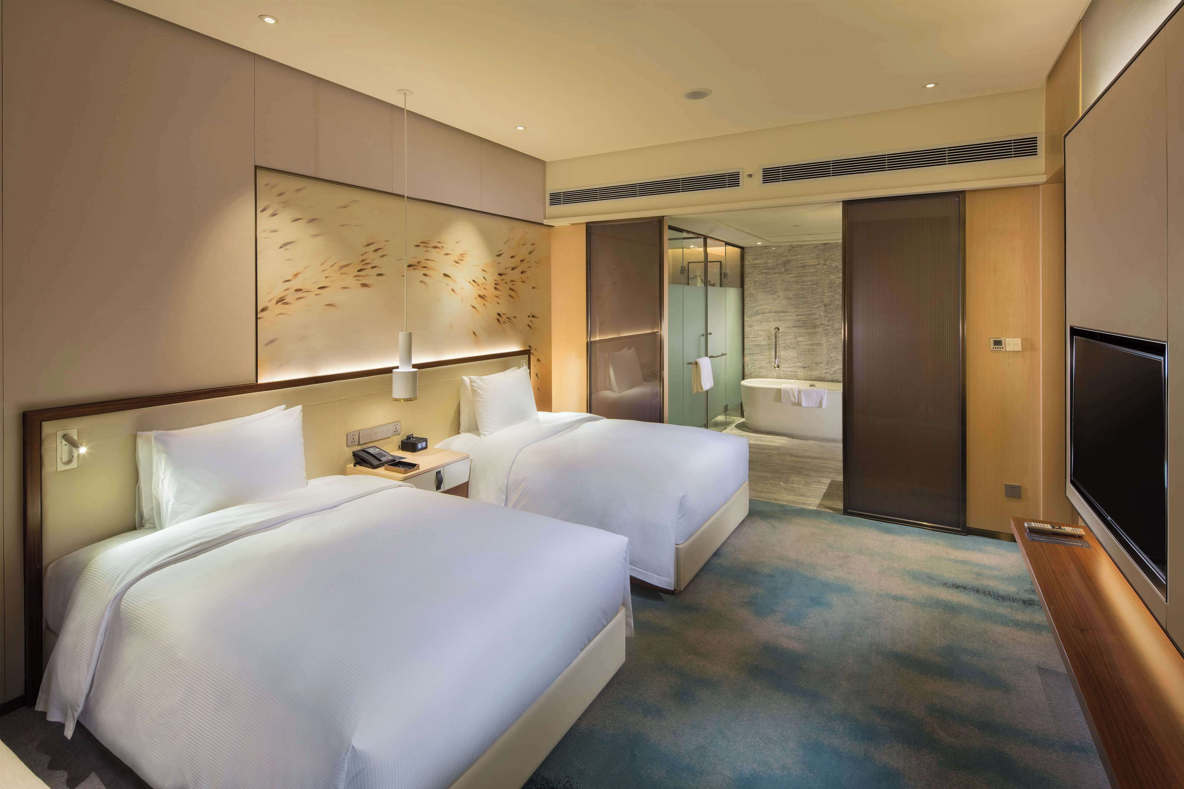 Habitación de hotel con dos camas, televisor, escritorio y baño visible a través de una puerta abierta.