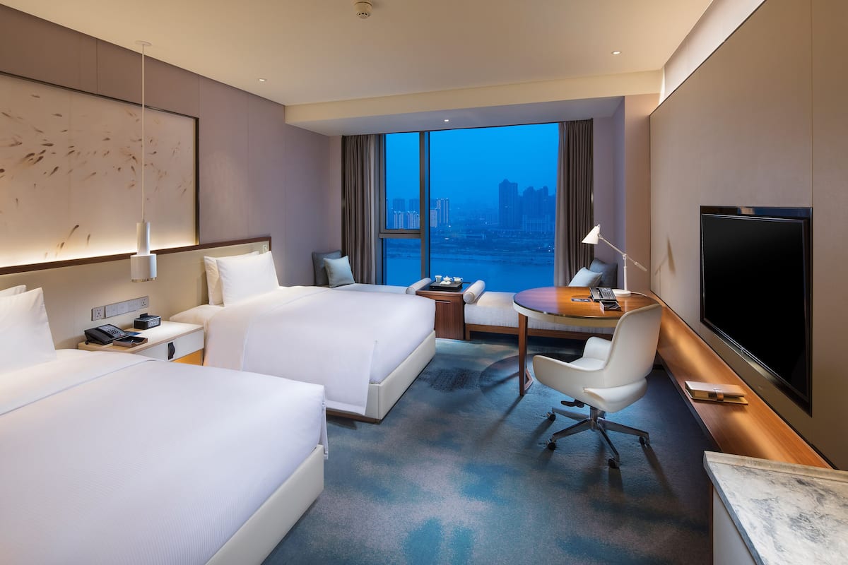 A hotel room with two beds, a desk, a chair, a TV, and a large window with a city view.