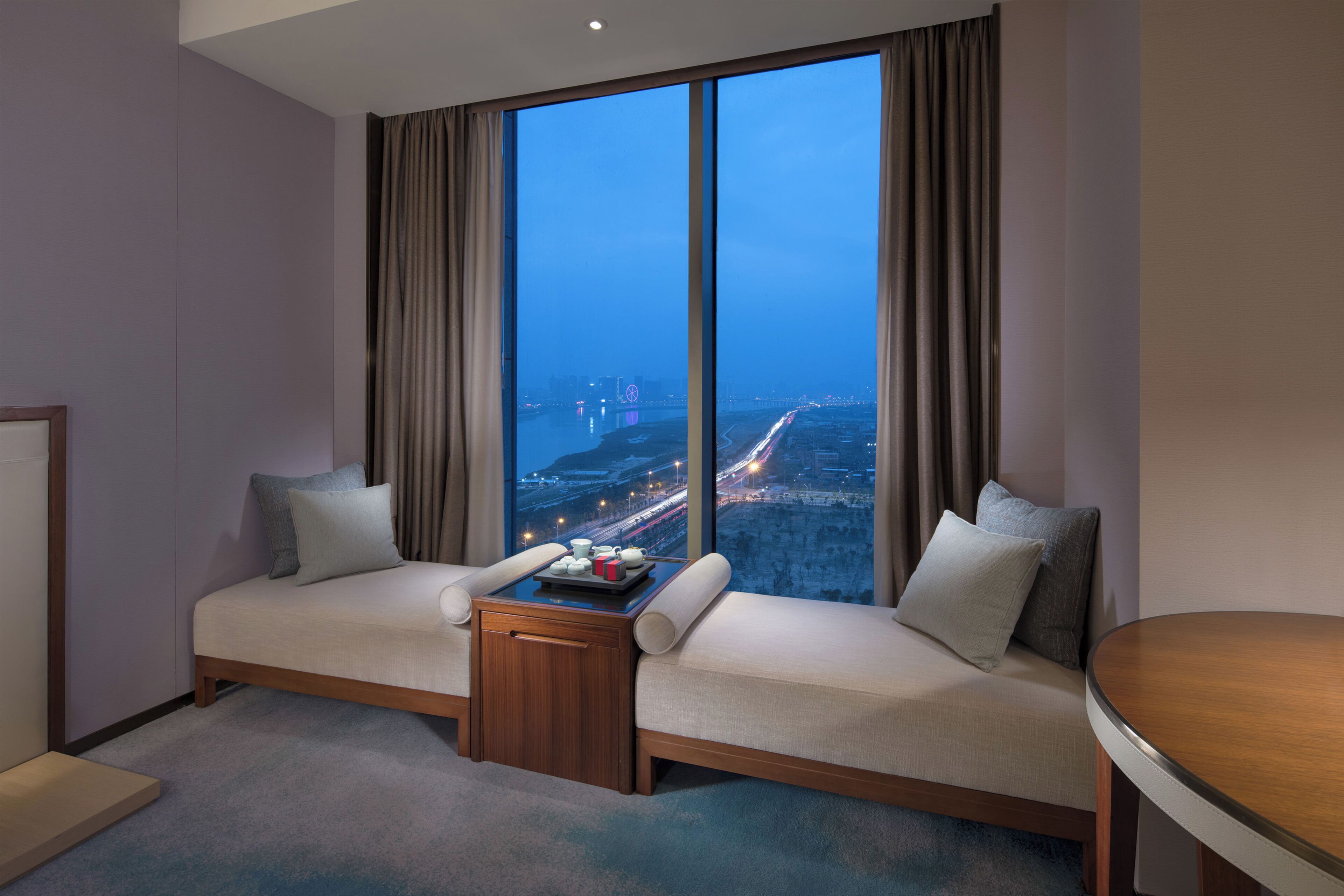 A hotel room with a large window, a sofa, a small table, and a view of a cityscape at night.
