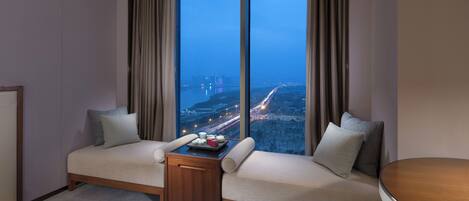 Habitación de hotel con un ventanal grande, un sofá, una mesita y vistas a un paisaje urbano nocturno.