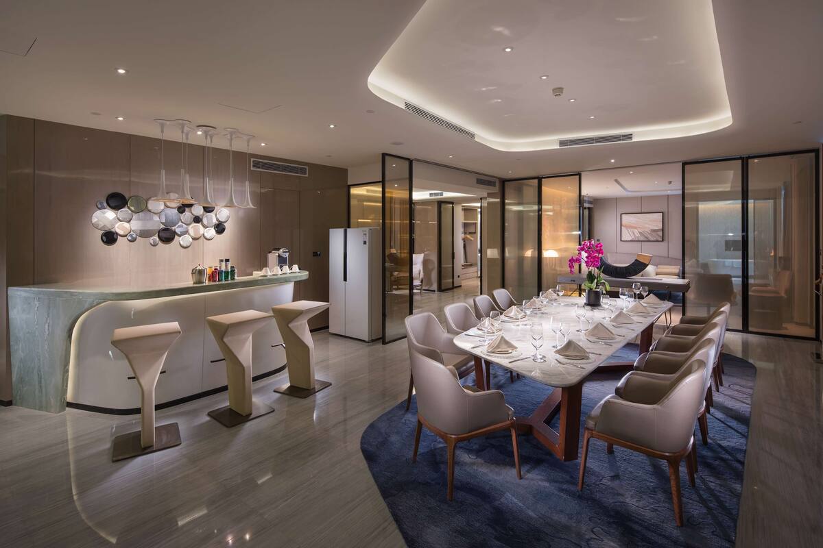 A modern dining area with a long table set for multiple guests, a bar with stools, and a contemporary light fixture above.