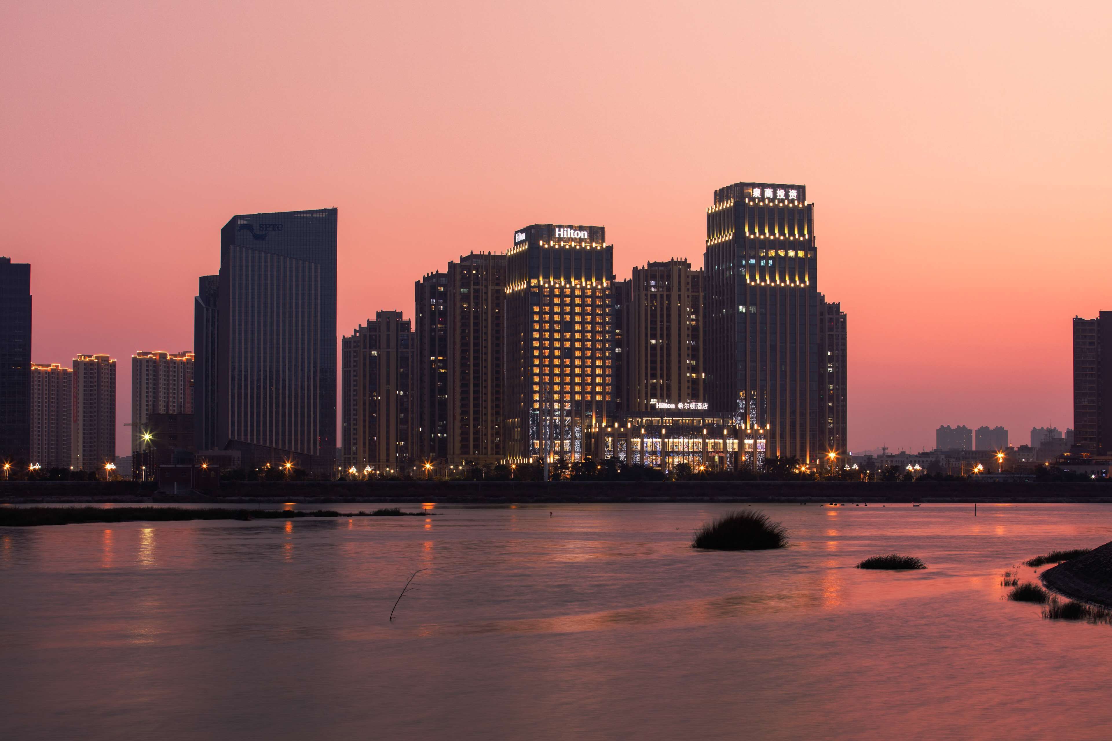 A city skyline with high-rise buildings, including a Hilton hotel, at dusk.