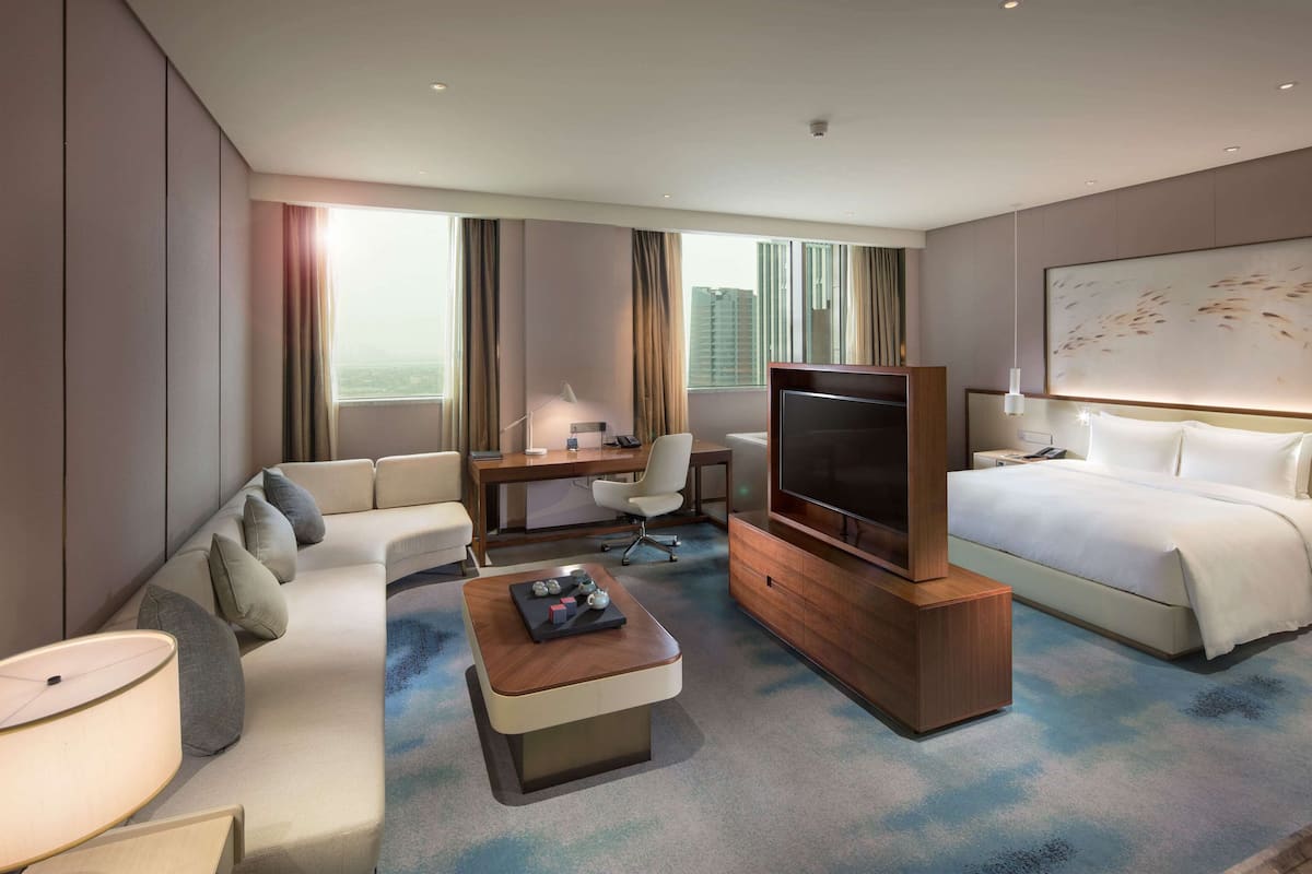 A modern hotel room with a sofa, a desk, a television, a bed, and a large window with curtains.
