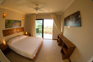 A bedroom with a large bed, a ceiling fan, a balcony with a view, and artwork on the walls.