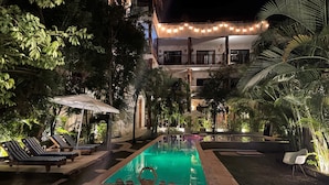 A nighttime pool area with lounge chairs and umbrellas, surrounded by lush greenery and a building with string lights. - Casa Tulum (Tulum)