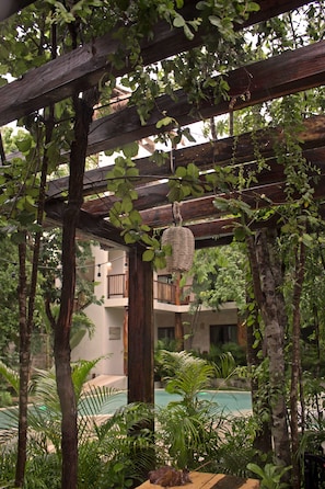 A wooden pergola with hanging wicker baskets, surrounded by lush greenery and a pool area.