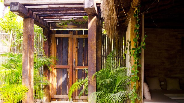 A rustic wooden structure with a thatched roof, a wooden door, and a bench with cushions. - Casa Tulum (Tulum)