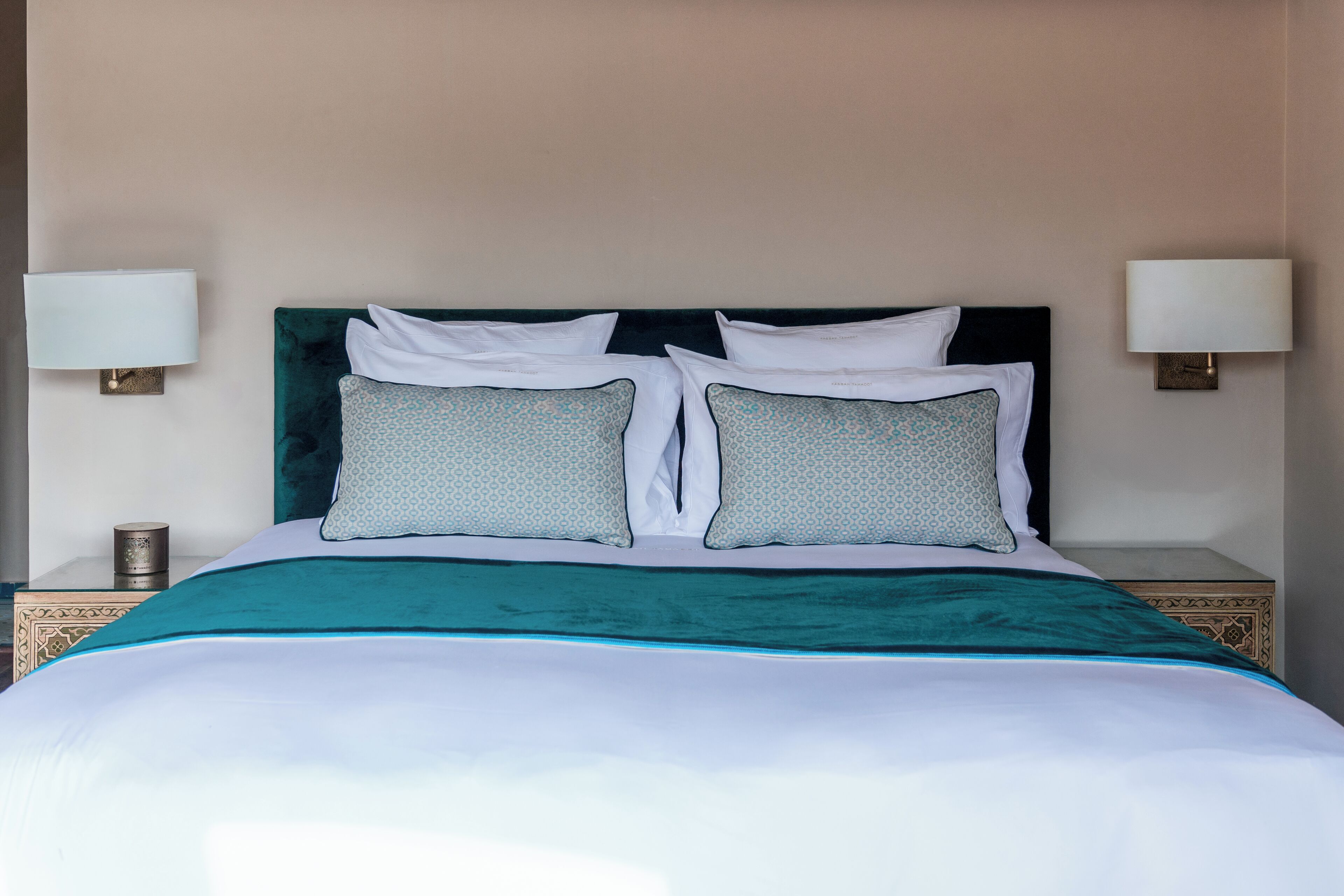A neatly made bed with a teal bedspread and two patterned pillows, flanked by two bedside lamps on matching nightstands.