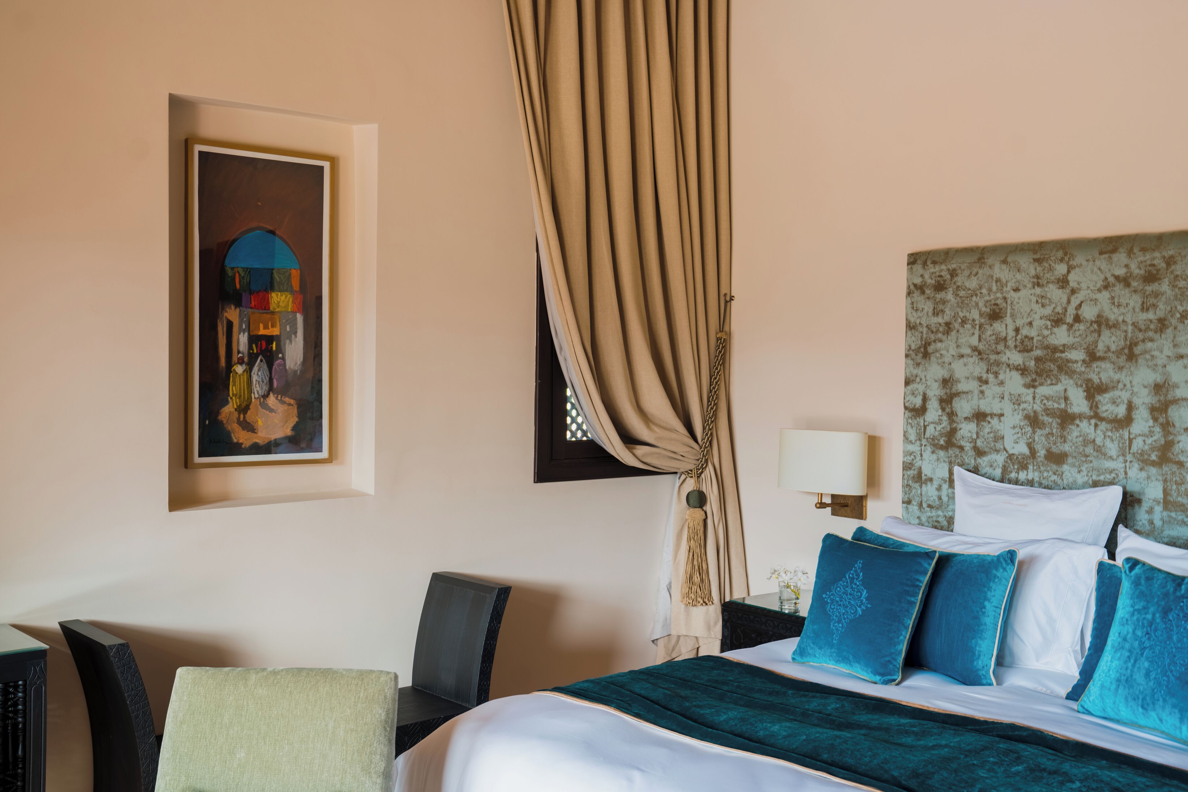 A hotel room with a bed, a chair, a desk, a lamp, and a painting on the wall.