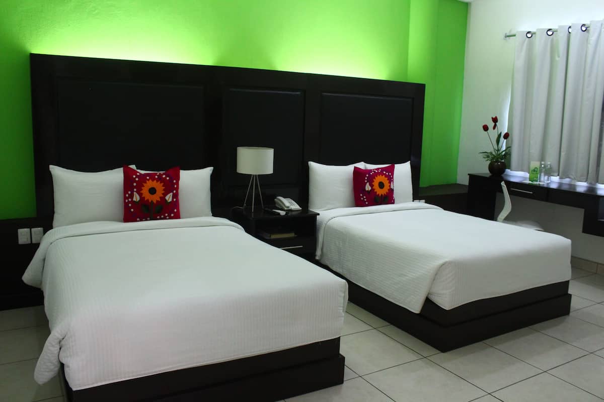A hotel room with two beds, a nightstand with a lamp, a window with curtains, and a vase with flowers.