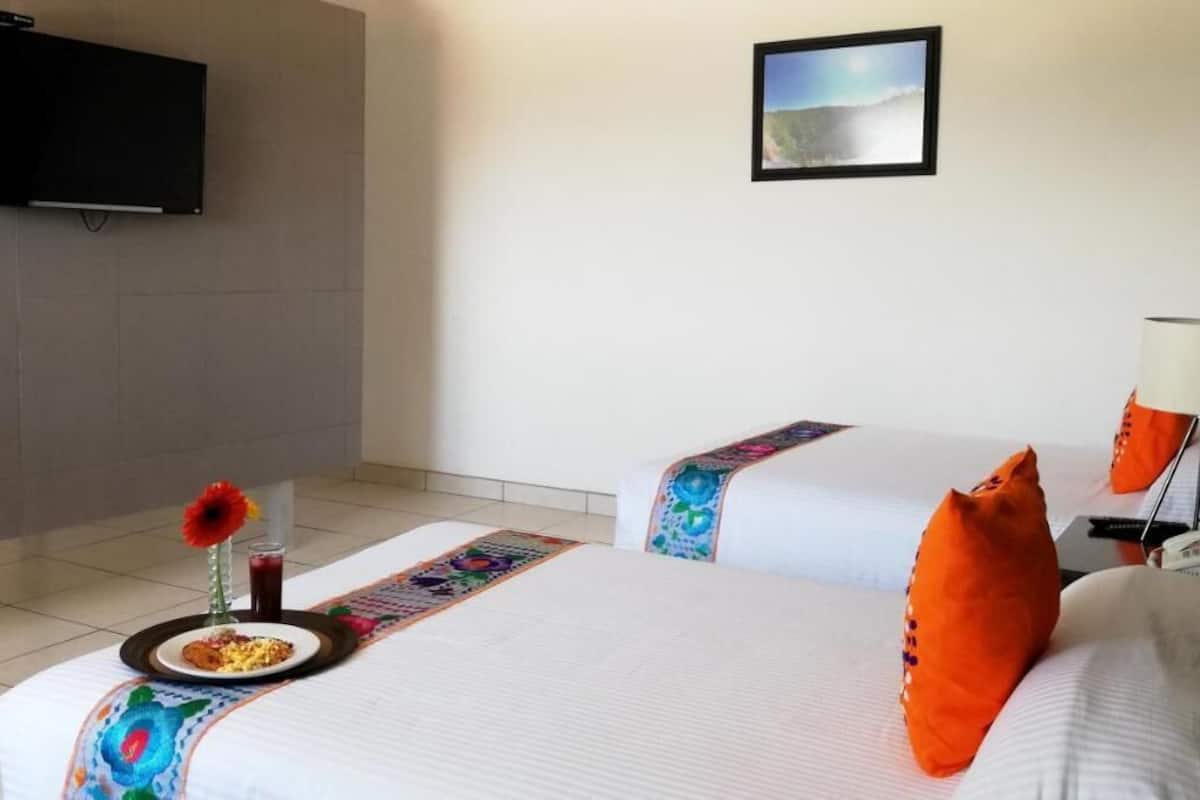 A hotel room with two beds, a flat-screen TV, a framed picture on the wall, and a small table with a plate of food and a drink.