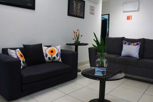 Una sala moderna con dos sofás oscuros, uno con un cojín floral y el otro con un diseño de sol. En el centro, una pequeña mesa redonda con un jarrón de plantas verdes y una revista.