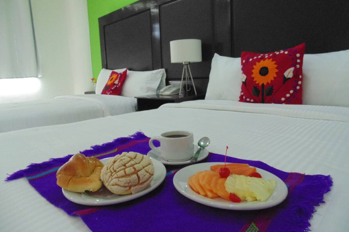 A hotel room with two beds, a coffee table with pastries, and a nightstand with a lamp.