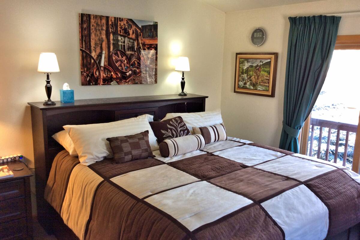 A neatly made bed with a brown and white quilt, two bedside lamps, a clock, and a painting on the wall.