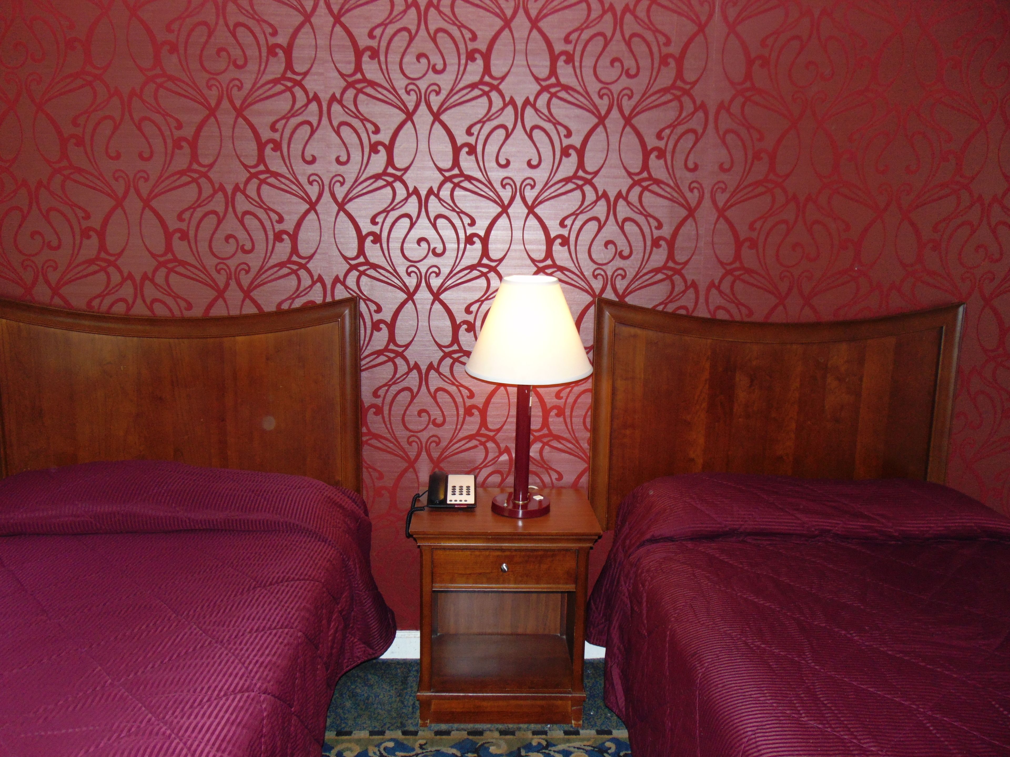 Habitación de hotel con dos camas, una mesita de noche con lámpara y un papel tapiz estampado.