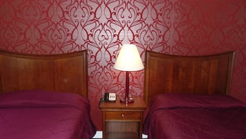 Habitación de hotel con dos camas, una mesita de noche con lámpara y un papel tapiz estampado.