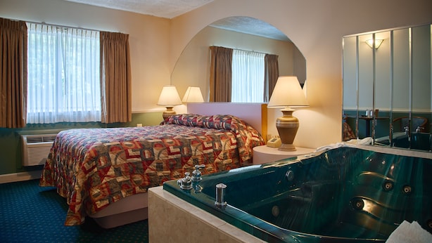 A hotel room with a large bed, a sitting area with a chair and a table, a large mirror, a bathtub, and a window with curtains. - La Mirage Motor Inn (Kendall Park)