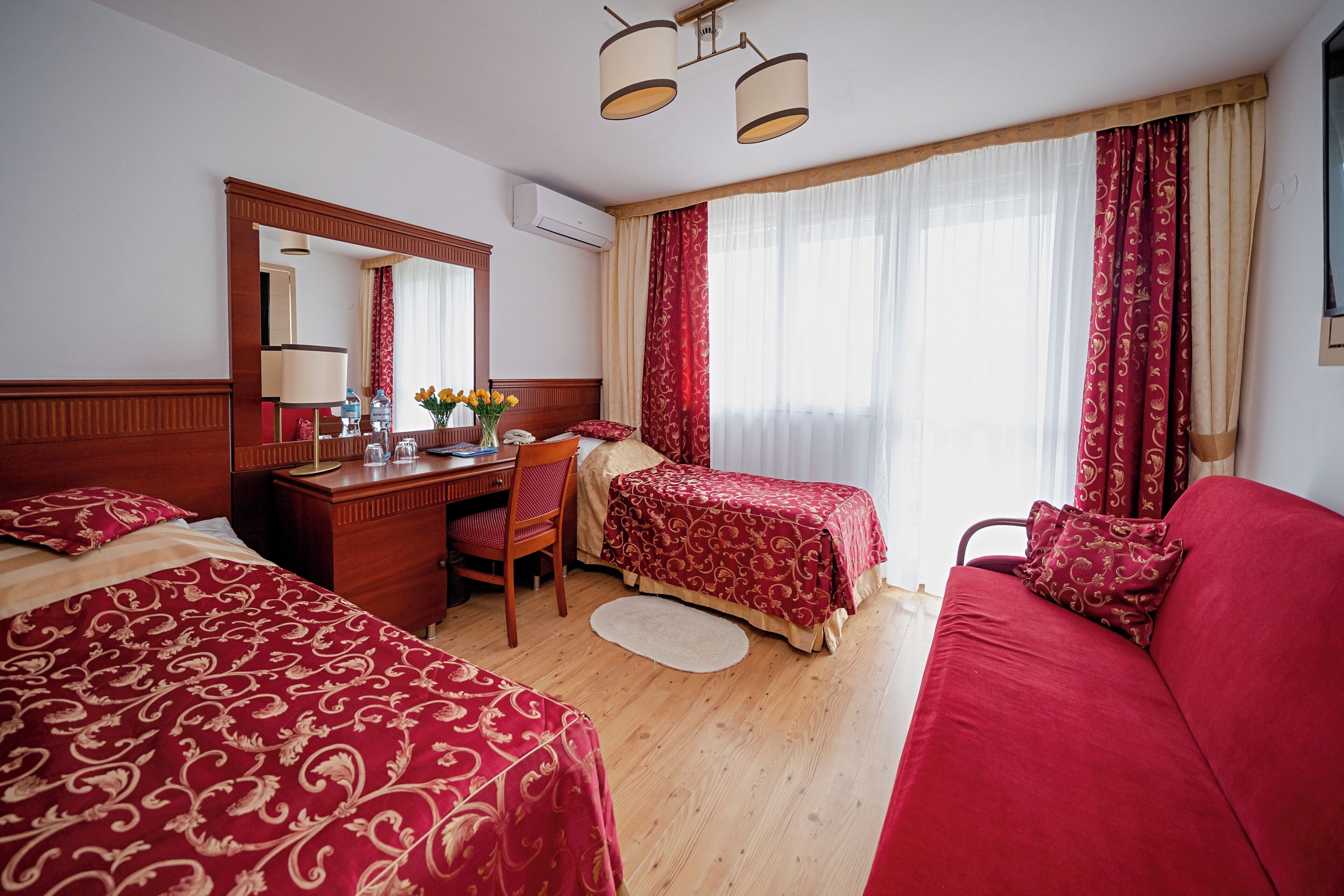 Een hotelkamer met twee bedden, een bureau, een stoel, een bank en een groot raam met gordijnen.