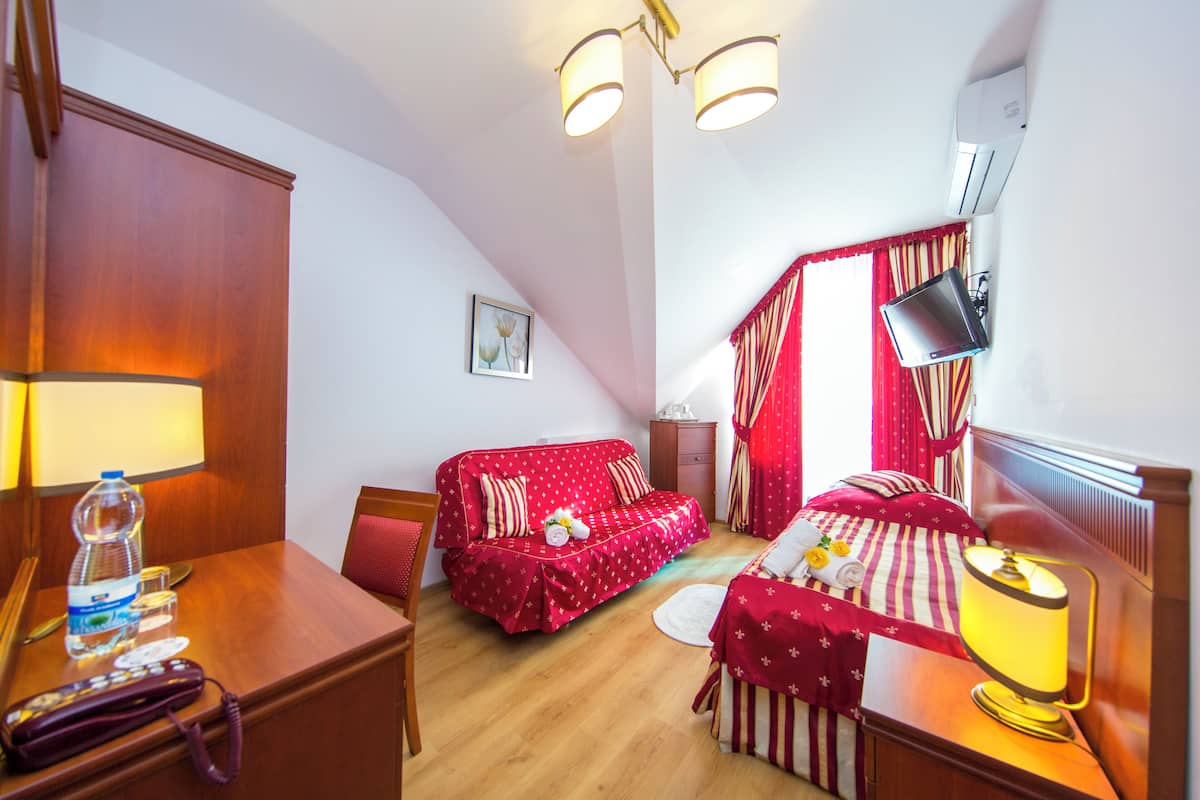 Una habitación de hotel con un escritorio de madera, un sofá rojo, una cama con ropa de cama a rayas, un televisor montado en la pared y una cortina roja.