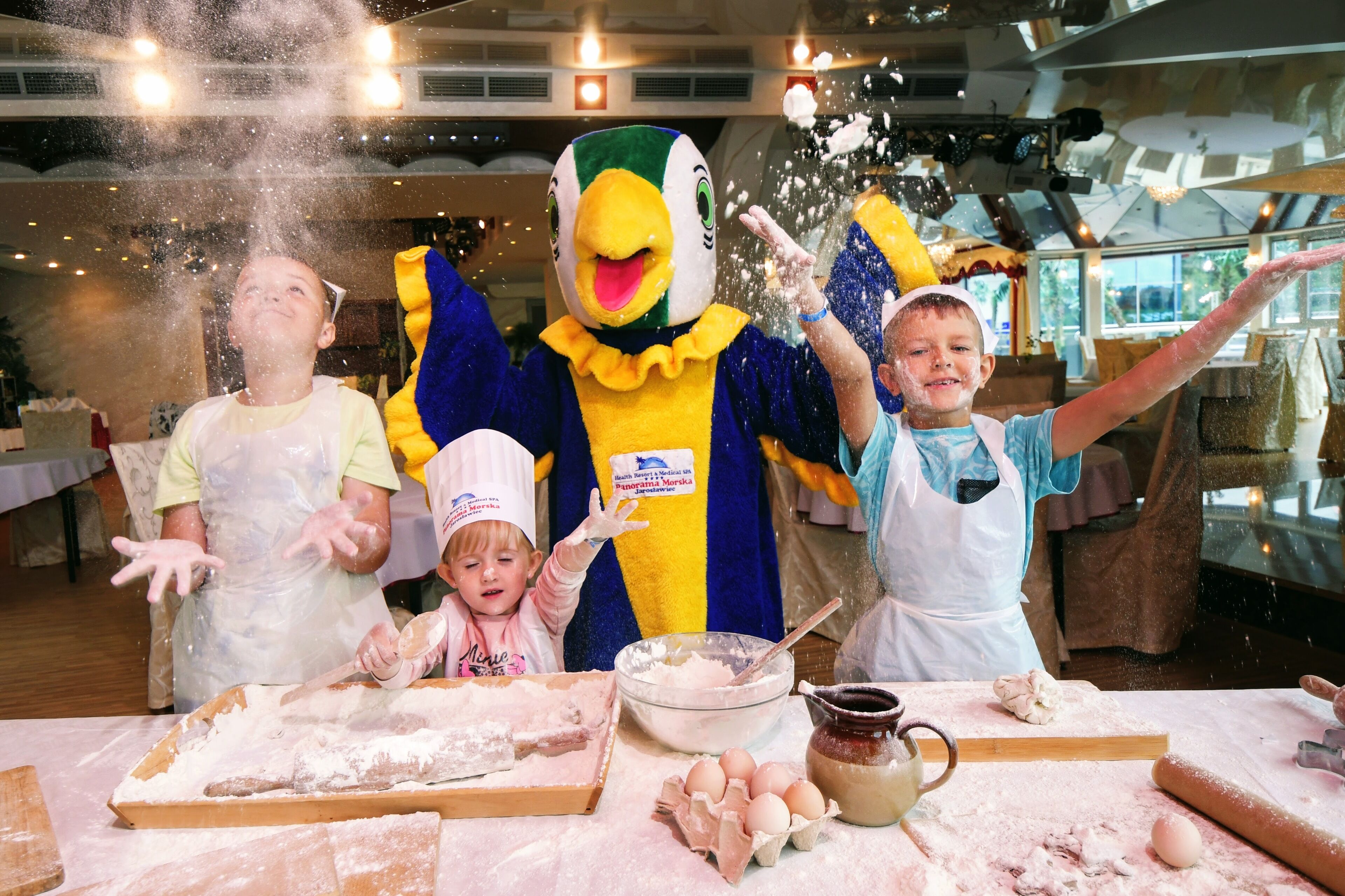 A person in a duck costume at a table with flour, dough, and eggs, suggesting a cooking activity.