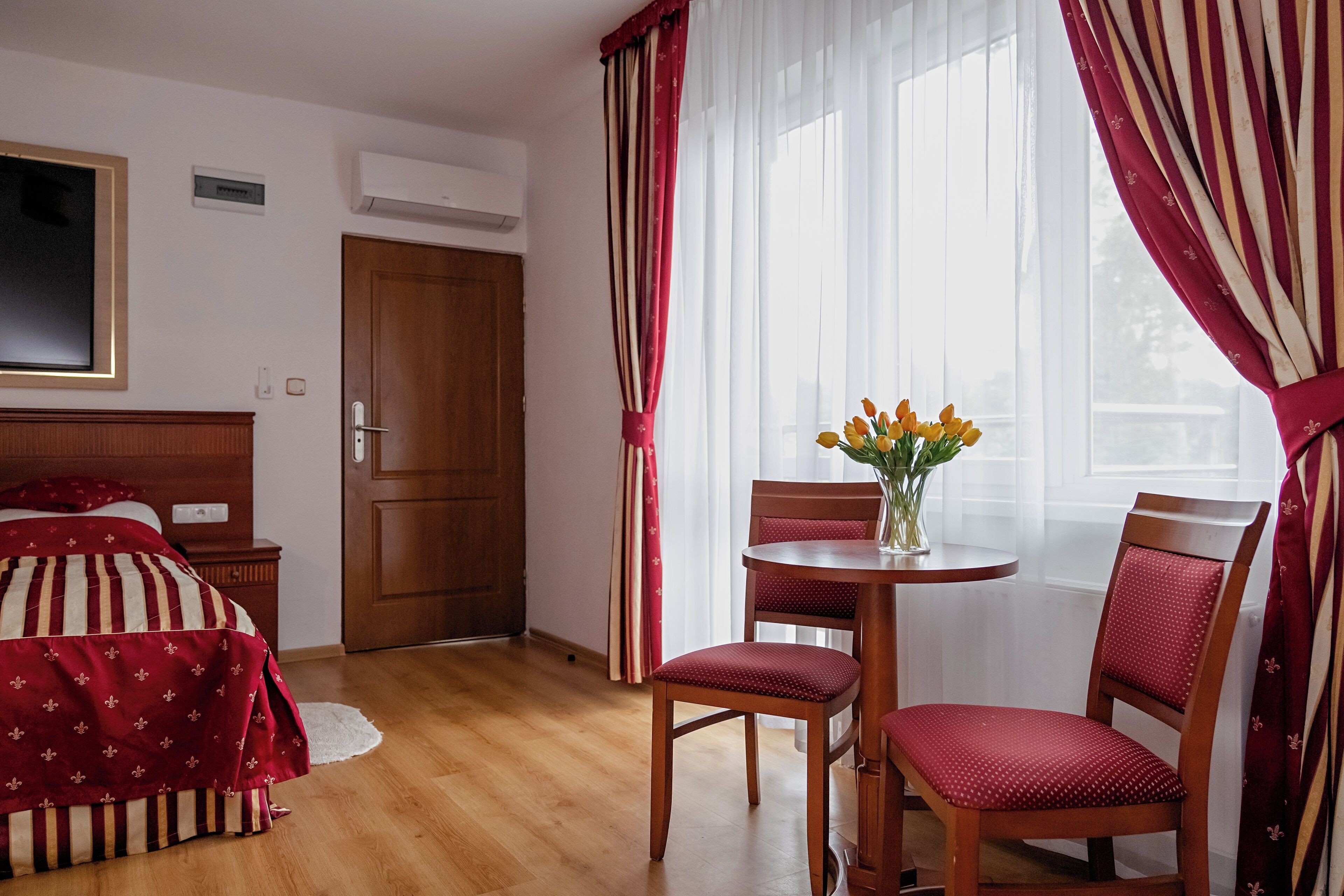 Habitació d'hotel amb un llit, una nit de fusta, una taula rodona amb un ram de flors, dues cadires vermelles i una porta de fusta.