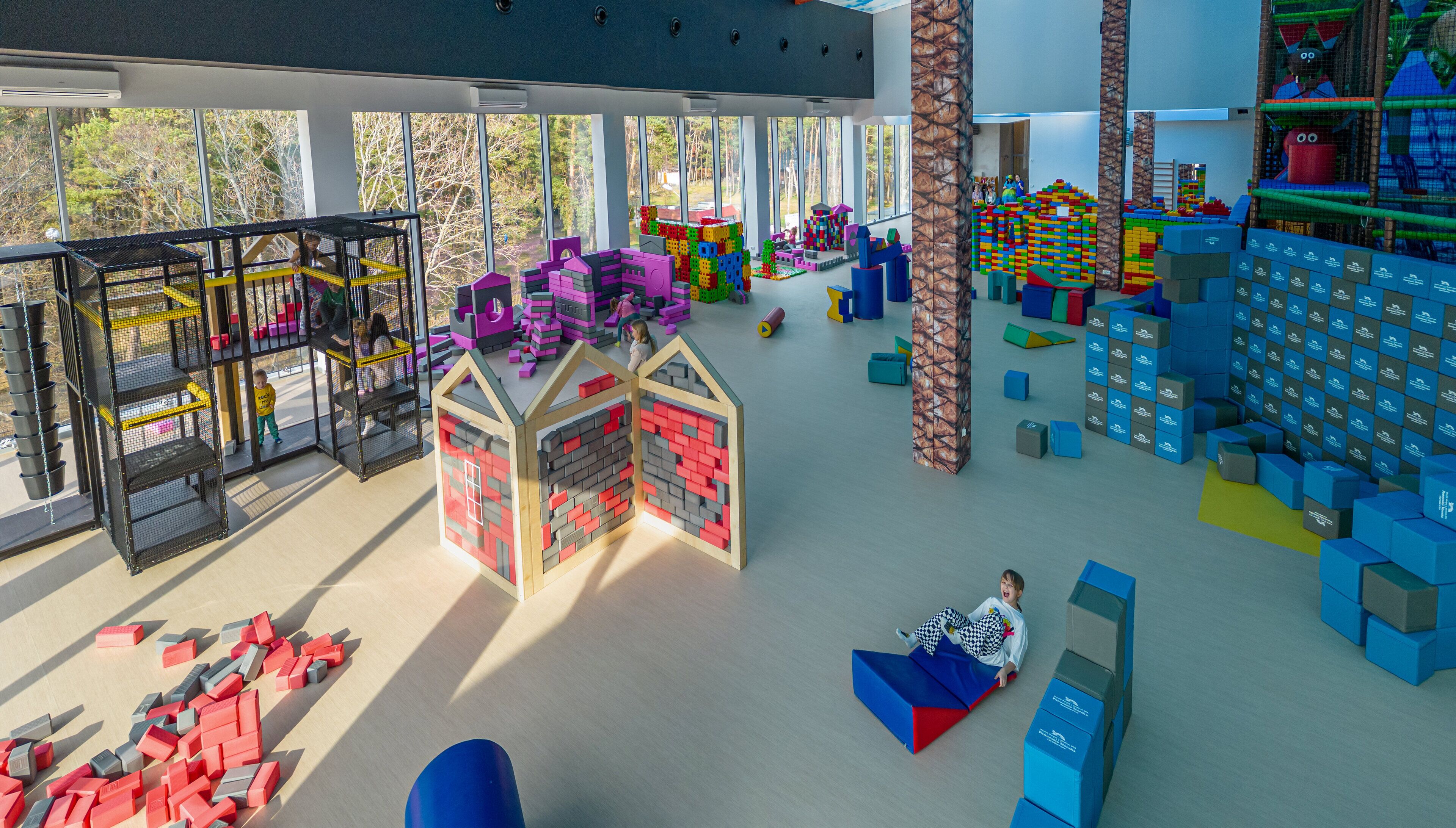 A spacious indoor play area with various play structures, including a climbing net, a house made of blocks, and a large climbing wall.